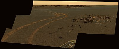 Figure 9. Sol 1162 (2007-05-01). The vicinity of the rim of Victoria Crater. This image shows a tiny (~1 m diameter) crater, Granada Crater,[44] and this crater's ejecta in the right foreground, and a large 800m diameter crater, Victoria Crater, in the left background. The tracks of the rover Opportunity wind across the image around the two craters. The larger crater is much older (> 140 million years) than the smaller one (a few hundred thousand years) and in a more advanced state of erosion.[24] An expanse of smooth sandy soils fill out the background. Image taken on Sol 1162 (2007-05-01).