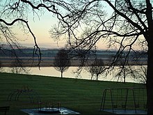 A children's play area sits amid leafless trees in the foreground. Beyond is a still body of water reflecting a cloudless sky. In the middle distance is a brown-coloured marshland, with green fields and woodlands beyond that, illuminated by a sun that is low in the sky.