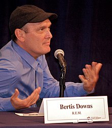 Bertis Downs looking to the camera's left and gesturing with his hands as he sits at a table behind a placard with his name