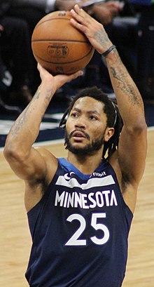 Hip length view of man raising the basketball to shoot free throw, with black moustache, beard, and short dreadlocks, thin white headband, wearing navy blue detroit uniform