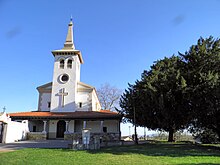 A picture of a church. The church is off to the left and is flanked on the right by some trees. There is a large cross above the door of the church and another cross at the very top of the bell tower.