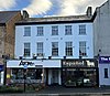 A white, rendered brick building on three storeys, in a terrace, seen from the north. The lower floor of the building is covered by shop fronts.