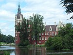 A red, ornate neo-gothic castle in a park-like location, the main tower of the castle is located to the left and topped with an ornate round dome and spire