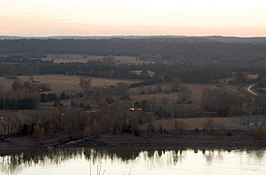 Looking across the Arkansas River from an overlook on Reed Mountain, the flat farmlands of the Arkansas River Valley stands in stark contrast to the mountain itself. Trees, roads, and homes are scattered along the river bottoms, and elevation slowly rises in the distance. Far in the background are the outlines of mountains in the orange-red sunset-colored sky