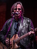 Musician Jackson Browne, playing an electric guitar and singing into a microphone.