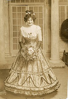 Phtographie sépia d'une drag queen en maquillage sophistiqué, portant des fleurs blanches dans ses cheveux bruns bouclés, un collier ras-de-cou avec une croix en pendentif, une robe longue corsetée à flots échancrée de dentelle blanche, des gants mi-longs de dentelle noire et tenant un bouquet de roses blanches et d'autres petites fleurs