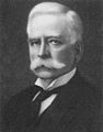 A headshot of a Caucasian male with a white handlebar moustache and white hair. He is wearing a white winged-collar shirt, black cravat and black coat.