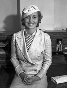 Martha Tilton sits atop a desk, wearing a light suit with a striped blouse.