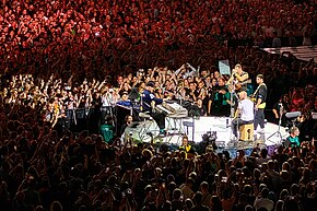 Four men perform on a small stage as a large crowd is gathered around them