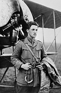 Half portrait of young dark-haired man in military uniform with coat over left arm, standing in front of biplane