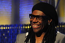 Photo d'un homme souriant avec des lunettes, un chapeau et une boucle d'oreille