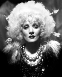 A grey-scale image of a woman wearing heavy make-up and a big, round, blond wig. She also wears garlands and feathers around her neck.