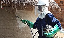 Person wearing personal protective equipment performing indoor residual spraying on a house to prevent mosquitoes.