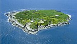 A small island, comprising a green plateau flanked by pale cliffs, with a lighthouse at the nearest point.