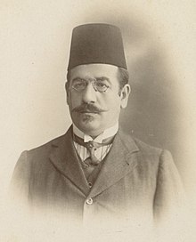 Portrait d'un homme moustachu portant un fez, un costume, une cravate et des lunettes.