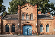 An ornate brick building with round arched barred windows, one story high except for a two-story high central entrance pavilion with a pointed roof. There are some tall shade trees behind it.