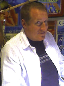 A bust photograph of a middle-aged caucasian man with short hair. He is wearing a white dress shirt over a black t-shirt. He is turned at a 45 degree angle away from the camera, but his eyes point directly at the photographer.