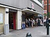 A modern looking building with marble coloured tiled wall at ground level with grey cladding above. A small concrete canopy is over the larger of two entrances with blue signs reading "SLOANE SQUARE STATION", people are entering and exiting through the larger entrance.