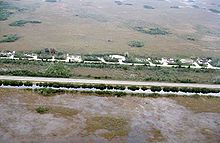 A color photograph taken from the air showing the Everglades bisected by a highway; at the bottom is a sawgrass field flooded with water bordered by a full canal; at the top are some homes and a dry sawgrass field