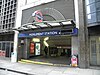 An entrance in a larger building under a sign reading "MONUMENT STATION" reveals banisters leading down. A woman is walking out of the entrance