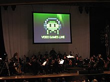 A photograph of an orchestra on a dimly lit stage. Above the group is a projection screen with a black, white, and green image of pixel art. The pixel art is of an oval object wearing headphones with eyes and four tentacles. Below the pixel art is the phrase "Video Games Live".