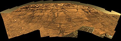 Figure 6. Burns Cliff in Endurance Crater. This image is one of the best known by Opportunity's Pancam. The original file is a huge (12,316 x 4,218 pixels) "mosaic" of many images. It has a "fish eye" view distortion. Click on the image for higher resolution. Burns Cliff is at least 7 m tall.[37] Meridiani sediment cliffs as big as Burns Cliff are not common. They only occur in large Meridiani craters younger than the sediments. The sediments in Burns Cliff are more weathered than those in Figure 5 since Endurance Crater is much older than Concepcion Crater.[24] Opportunity collected data at Burns Cliff over several months in 2004, and many important papers used this data.[37][38][39][40][41][42] Images taken on Sols 287-294 (2004-11-13 to 2004-11-20).