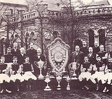 A sepia photograph with a large old structure in the background obscured by trees. In the foreground there is a large shield surrounded by five trophies. On either side of the shield stands 8 people