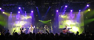 Four-man rock group performing on stage, spotlit, with row of darkened fans in the foreground