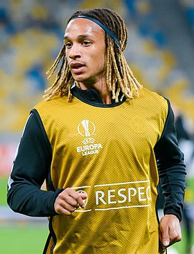 Photographie couleur d’un homme, de tronc, cheveux mi-longs façon dreadlocks décolorées tenus par un serre-tête bleu, portant une chasuble jaune par-dessus un pull foncé