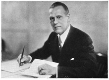 Black and white photograph of a man wearing suit holding a pen with one hand and a piece of paper with the other.