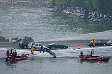 Emergency personnel running on the collapsed bridge and two Minneapolis Fire Department boats in the water with dozens of observers on the bank of the Mississippi