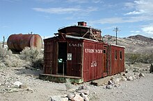 An abandoned wooden rail car rests on a bed of gravel. A large, rusting, cylindrical tank is nearby, among low bushes. Barren, striated hills can be seen in the distance. Lettering on the car says, "L.A. & SL" and "Union Pacific". A sign on the end of the car says, "Danger: Do Not Enter" and "Peligro: No Entre