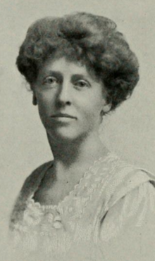 A white woman with hair in a bouffant style, wearing a white lace-trimmed top or dress