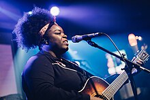 Une femme noire bouche ouverte devant un micro. Elle tient une guitare.