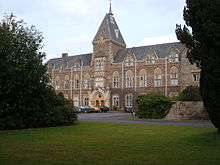 Red brick building with tower. In the foreground are grass and shrubs.