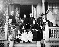 Late 19th century photo of a family of ten adults and four children posing on the front porch of a large house. The women wear long dresses with high collars and leg-o-mutton sleeves. The men have flowing moustaches. Marjory is a toddler in a long dress and black stockings.