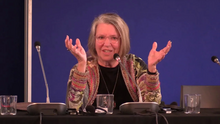 Nancy Fraser, mit grauen Haaren und Brille sitzt hinter einem Mikrofon auf einem Podium.