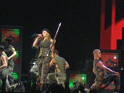 Image shows a performance onstage where the performers are wearing military garments. Central to them is a blond woman, who carries a rifle in her left hand while singing into a microphone.