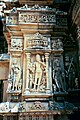 A fierce form of Devi, Chamunda is portrayed as a frightening, emaciated hag. Her attributes include fangs, a sword, Shiva's trident, skull garland, skull staff, skull bowl, and skeletal corpse which she tramples or sits on. Woman playing eka-tantri vina, far right.