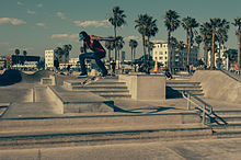 Photo d'un skater en train d'effectuer une figure de skate à Venice Beach. Il est pris en l'air durant un saut à 1 à 2 mètres de hauteur et son skate effectue des rotations en dessous de lui. Le décor est principalement composé d'un multitude d'escaliers en béton de quelques marches. En arrière-plan assez loin, il y a des skaters et beaucoup plus loin, on aperçoit des cocotiers, la plages et des immeubles.