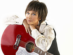 A woman with short brown hair, wearing a white shirt and leaning on a red guitar