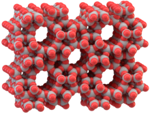 Eine Computergrafik aus grauen und roten Kugeln. Die Kugeln sind teilweise ineinander geschoben und bauen so eine räumliche Struktur auf. Graue Kugeln sind paarweise durch jeweils eine rote Kugel verbunden. In der Struktur sind fünf zylindrische Hohlräume, um die herum sich das Netzwerk der Kugeln legt. Die Hohlräume sind untereinander symmetrisch angeordnet und ihr Durchmesser ist in etwa genau so breit wie der Abstand zwischen zwei Hohlräumen.