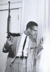 A man in a suit and tie, carrying a rifle, looks out the window