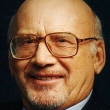 Close-up, full-face publicity photograph of a smiling, elderly man with spectacles, designer stubble, and receding white hair.