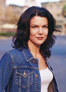 A young brunette woman smiling towards the camera, wearing a denim jacket over a white tank top.