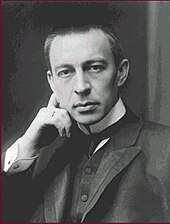 A clean-shaven young man with close-cropped hair, wearing a dark jacket, dress shirt with a high stiff collar and tie, looking toward the viewer with one hand on the side of his head