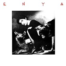 A black and white photograph of a brunette woman with a dark dress and high heels, crouched down, with two aggressive stuffed dogs beside her. The image is surrounded by a white border and the word "Enya", stylized in all capital red letters, is placed on the top corner of the picture.